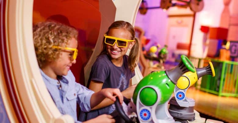 Two smiling children wearing yellow 3D glasses play with toy cannons at an indoor Disney's Hollywood Studios attraction, surrounded by colorful decorations and a playful atmosphere.