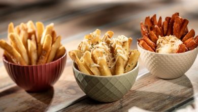 Three bowls of fries on a wooden surface: classic fries in a red bowl, cheese and herb-topped fries in a green bowl, and seasoned sweet potato fries with dip in a white bowl.