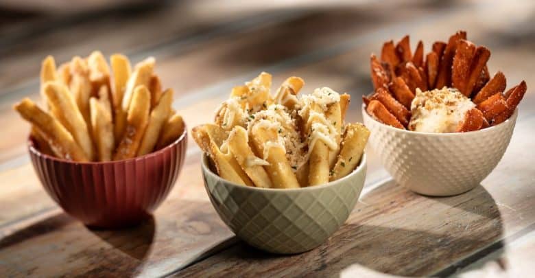 Three bowls of fries on a wooden surface: classic fries in a red bowl, cheese and herb-topped fries in a green bowl, and seasoned sweet potato fries with dip in a white bowl.