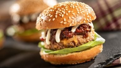 A close-up of a juicy hamburger in a sesame seed bun with lettuce, a beef patty, melted cheese, sauce, and relish, set on a dark surface with another burger and a plaid cloth in the background.