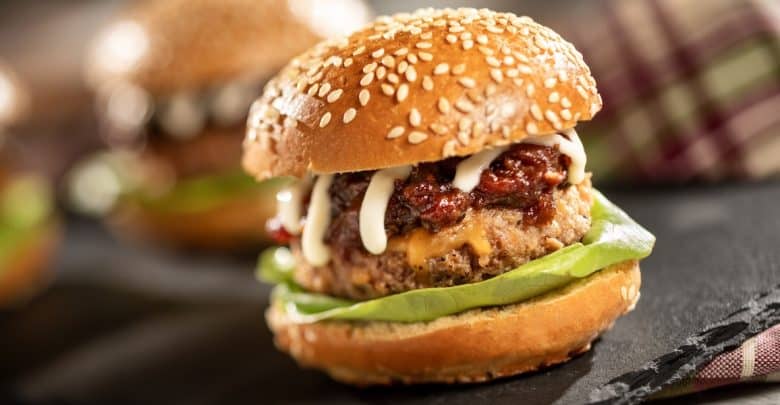 A close-up of a juicy hamburger in a sesame seed bun with lettuce, a beef patty, melted cheese, sauce, and relish, set on a dark surface with another burger and a plaid cloth in the background.