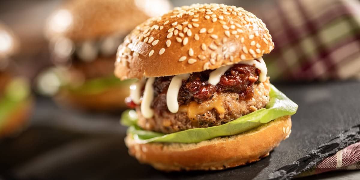 A close-up of a juicy hamburger in a sesame seed bun with lettuce, a beef patty, melted cheese, sauce, and relish, set on a dark surface with another burger and a plaid cloth in the background.