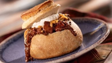A round bread bowl filled with chili, topped with shredded cheese and sour cream, with the cut-out bread lid resting on top. The chili is spilling over the side, and the bowl is on a blue plate.
