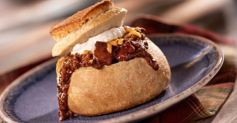 A round bread bowl filled with chili, topped with shredded cheese and sour cream, with the cut-out bread lid resting on top. The chili is spilling over the side, and the bowl is on a blue plate.