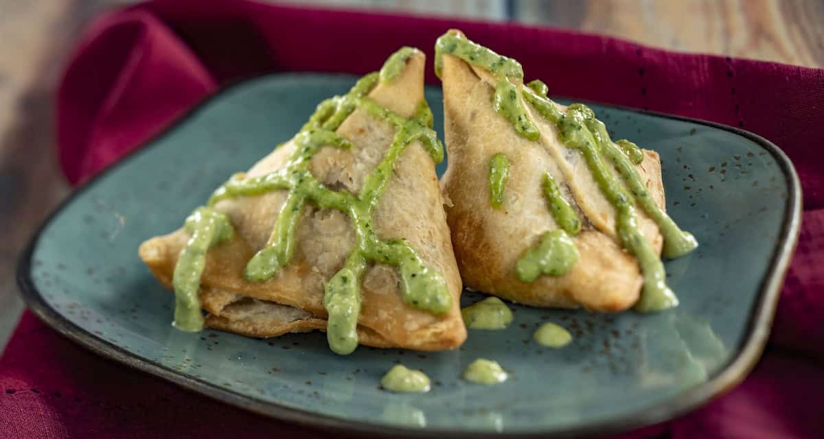 Two golden-brown, triangular samosas topped with green chutney are served on a blue plate with a maroon cloth in the background.