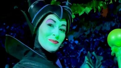 Maleficent with a black horned headpiece, and a glowing staff, smiling under dramatic green and blue lighting.