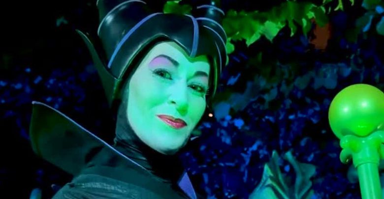 Maleficent with a black horned headpiece, and a glowing staff, smiling under dramatic green and blue lighting.