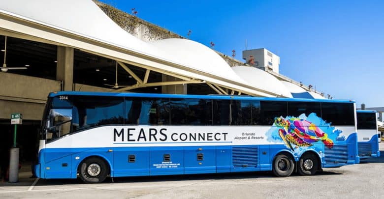A blue and white Mears Connect bus, with colorful graphics and "Orlando Airport & Resorts" written on the side, is parked outside a modern building under a clear blue sky.