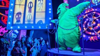 A performer in a large, bright green Oogie Boogie costume greets an excited crowd of children in a vibrant, spooky setting with colorful, glowing decorations and animated skeleton images in the background.