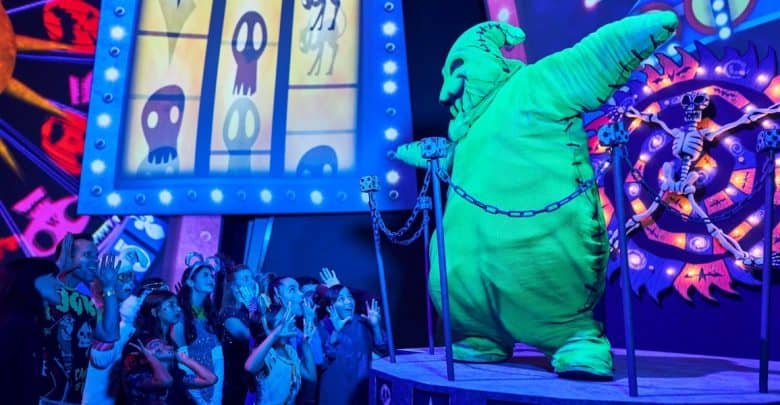 A performer in a large, bright green Oogie Boogie costume greets an excited crowd of children in a vibrant, spooky setting with colorful, glowing decorations and animated skeleton images in the background.