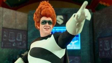 A person in a black-and-white superhero costume with a large "S" on the chest and wild red hair points forward, standing in front of futuristic screens and equipment.