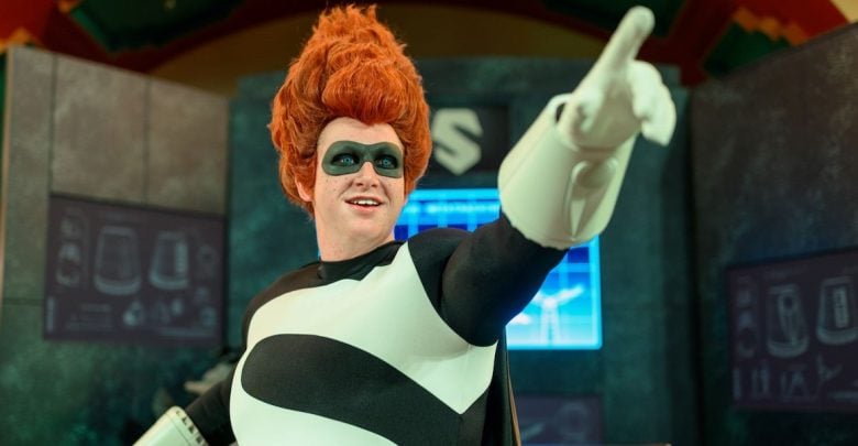 A person in a black-and-white superhero costume with a large "S" on the chest and wild red hair points forward, standing in front of futuristic screens and equipment.