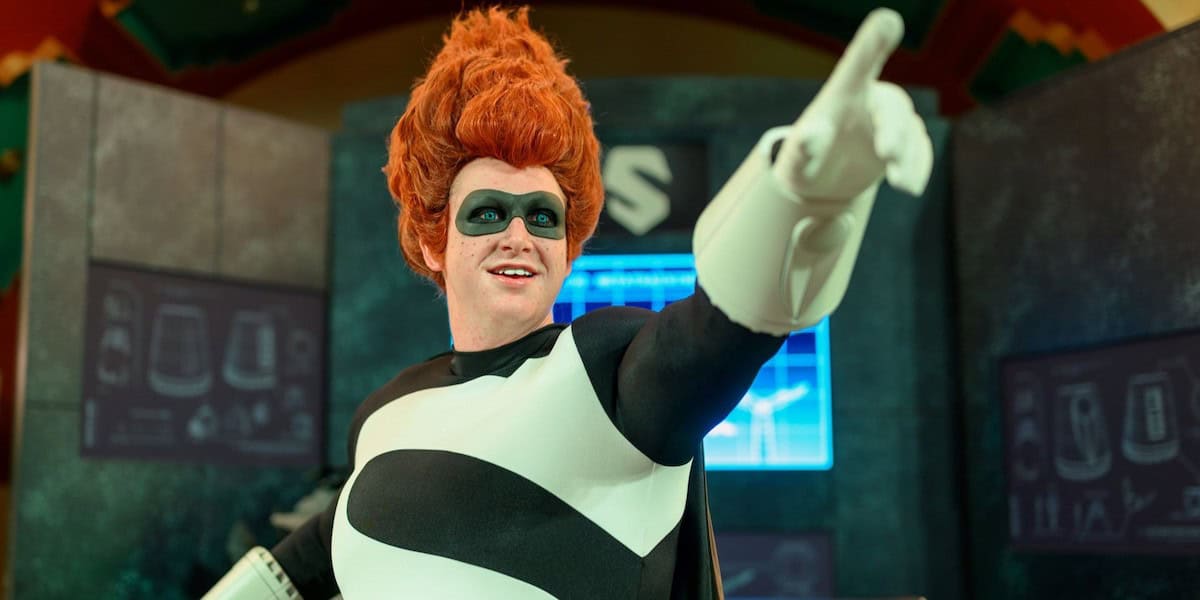 A person in a black-and-white superhero costume with a large "S" on the chest and wild red hair points forward, standing in front of futuristic screens and equipment.