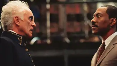 Two men stand facing each other indoors; one older with white hair wearing a dark uniform, and one younger with a mustache in a tan suit, appearing serious and engaged in conversation.