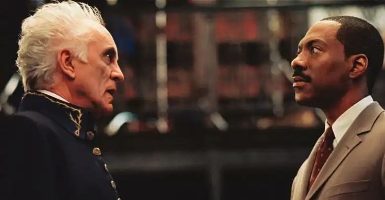 Two men stand facing each other indoors; one older with white hair wearing a dark uniform, and one younger with a mustache in a tan suit, appearing serious and engaged in conversation.