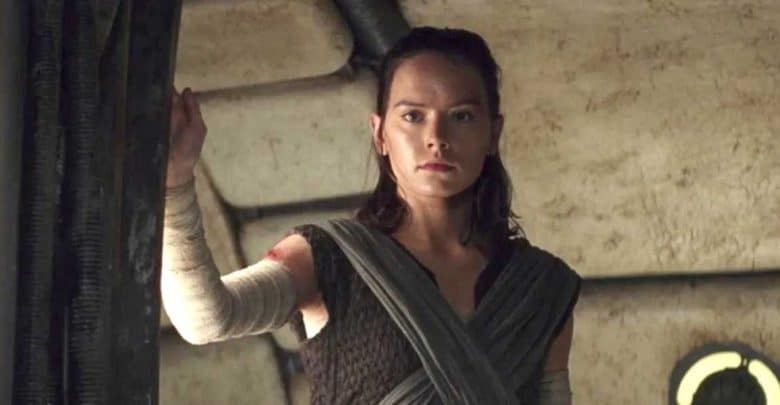 A woman with brown hair stands indoors, wearing a dark, textured outfit with bandaged arms. She looks serious and determined, holding onto a metal doorway in a sci-fi setting.