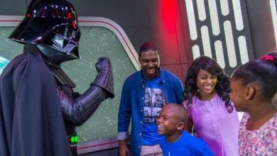 A family smiles and laughs while meeting a person dressed as Darth Vader in a sci-fi themed setting with a planet visible in the background.