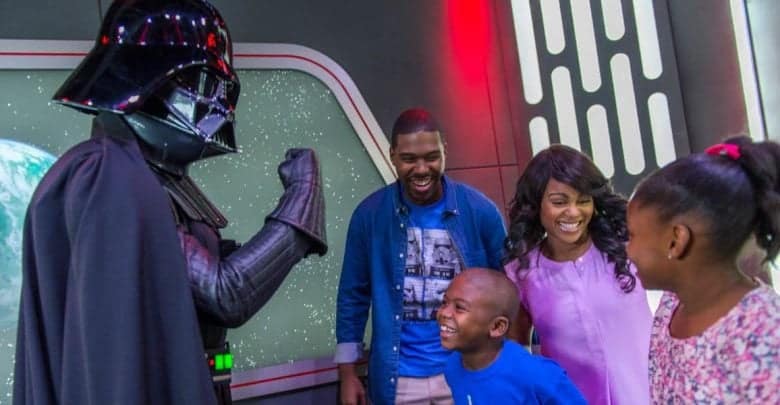 A family smiles and laughs while meeting a person dressed as Darth Vader in a sci-fi themed setting with a planet visible in the background.