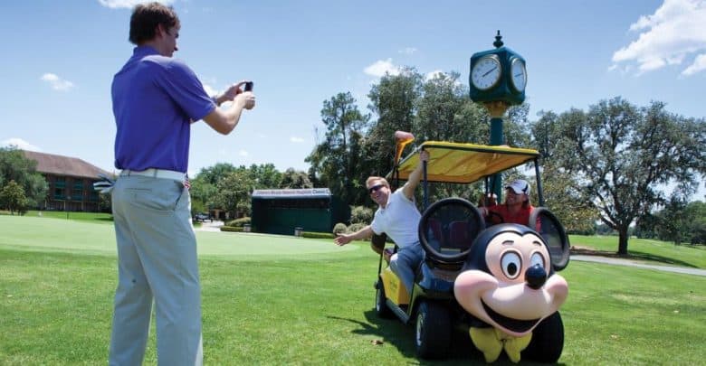 Two people pose in a golf cart decorated with a large Mickey Mouse face while another person takes their photo on a sunny golf course with green grass and trees in the background.