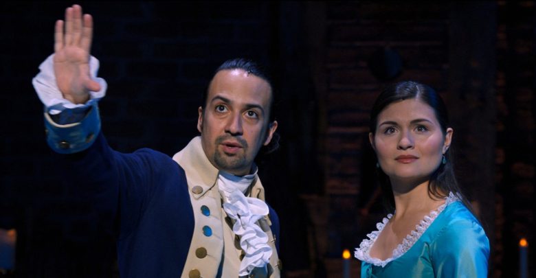 A man in 18th-century attire raises his hand while standing beside a woman in a teal dress with lace trim; both look ahead on a dimly lit stage with a dark background.