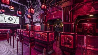 A futuristic, neon-lit room with red and purple lighting, displaying glowing panels and monitors. Roped barriers guide visitors through the space. Industrial and sci-fi design elements dominate the setting.