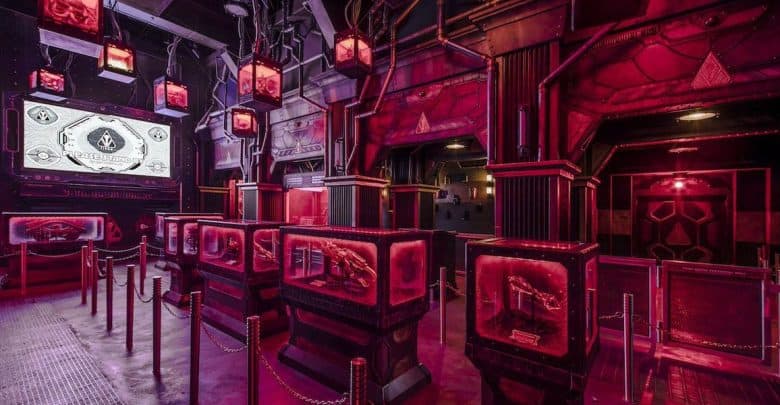 A futuristic, neon-lit room with red and purple lighting, displaying glowing panels and monitors. Roped barriers guide visitors through the space. Industrial and sci-fi design elements dominate the setting.