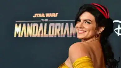 A woman in a yellow off-the-shoulder dress smiles over her shoulder on a red carpet in front of a "Star Wars: The Mandalorian" backdrop.