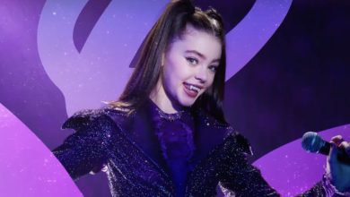 A girl in a sparkly purple outfit with high ponytails holds a microphone and smiles, revealing vampire fangs, against a purple background with swirling patterns.