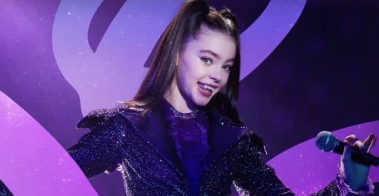 A girl in a sparkly purple outfit with high ponytails holds a microphone and smiles, revealing vampire fangs, against a purple background with swirling patterns.