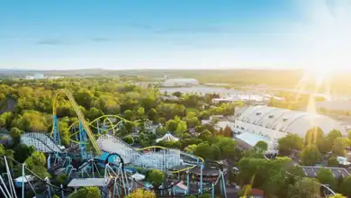 Aerial view of an amusement park with roller coasters, green trees, white domed buildings, and a bright sun shining in a clear blue sky.