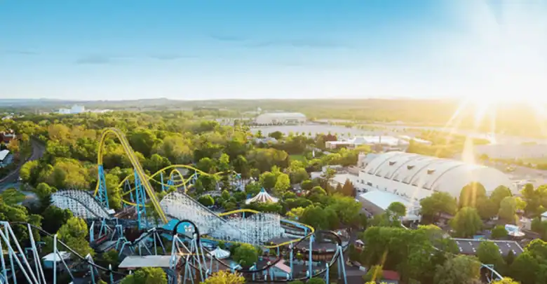 Aerial view of an amusement park with roller coasters, green trees, white domed buildings, and a bright sun shining in a clear blue sky.
