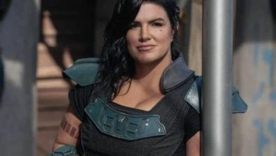A woman in futuristic armor with shoulder pads stands outdoors, leaning casually against a metal pole. She has long dark hair and a confident expression, with a tattoo visible on her right arm.