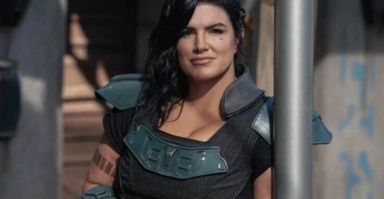 A woman in futuristic armor with shoulder pads stands outdoors, leaning casually against a metal pole. She has long dark hair and a confident expression, with a tattoo visible on her right arm.