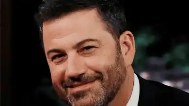 A man with short dark hair and a beard is smiling, wearing a dark suit and white shirt, with a blurred background.