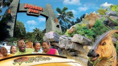 A group of people excitedly ride a yellow raft past a large dinosaur model at the Jurassic Park River Adventure attraction, with a Jurassic Park sign and lush greenery in the background.