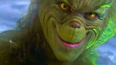 A close-up of the Grinch’s face, covered in green fur with yellow eyes and a mischievous smile, set against a snowy, bluish background.