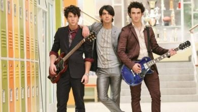 Three young men stand in a school hallway by yellow lockers, two holding guitars, all dressed in stylish suits. Trophies are visible on shelves in the background near a staircase.