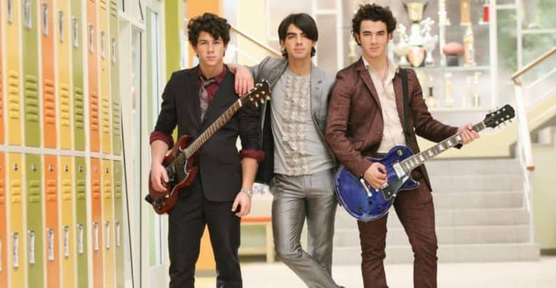 Three young men stand in a school hallway by yellow lockers, two holding guitars, all dressed in stylish suits. Trophies are visible on shelves in the background near a staircase.