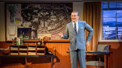 A man in a gray suit stands smiling in an office with wood-paneled walls, vintage decor, a large map, and photos on the wall. A window shows a blue sky outside. A desk and chair are beside him.