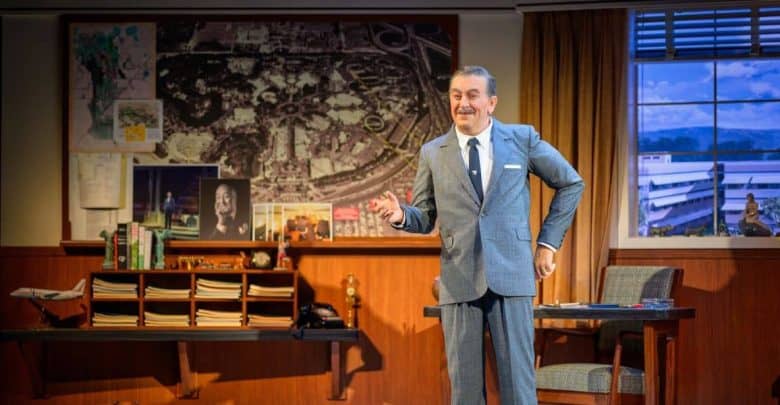 A man in a gray suit stands smiling in an office with wood-paneled walls, vintage decor, a large map, and photos on the wall. A window shows a blue sky outside. A desk and chair are beside him.