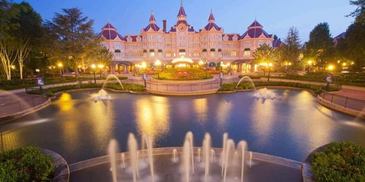 A grand, castle-like hotel with turrets and warm lights reflected in a large pond with fountains at dusk, surrounded by trees and manicured gardens.