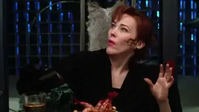 Catherine O'Hara lookalike with curly red hair and black gloves reacts in surprise at a themed restaurant table of food and drinks.