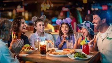 A group of six people of various ages enjoying a meal together at a restaurant. The kids are wearing Mickey Mouse ears, and everyone looks happy and engaged in conversation. The table is full of food, including pizza, burgers, and colorful drinks at a Disneyland park resort.