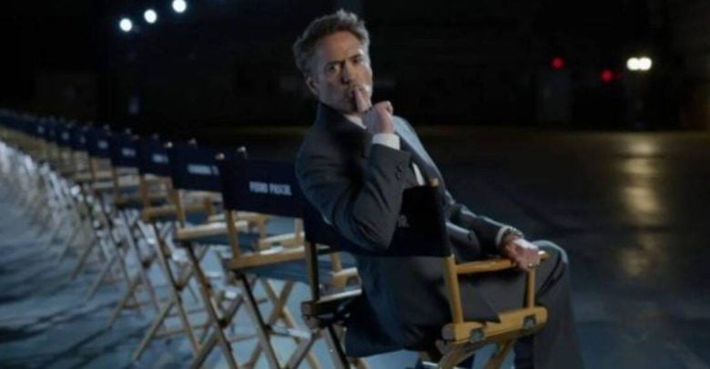 A suited man in a director’s chair on a dim Marvel Studios set, evoking Robert Downey Jr., with empty chairs behind him.