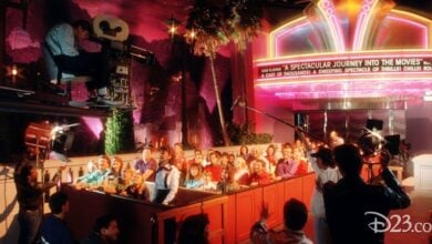 A film crew captures Disney park-level excitement as a crowd performs beneath a vibrant, retro movie theater marquee.