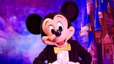 Mickey Mouse in a tuxedo with a yellow bow tie stands smiling before a colorful, magical backdrop featuring the iconic castle at Shanghai Disney Resort.