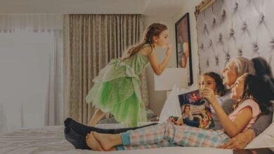 Three kids and their grandmother lounge on a bed, flipping through a Disney magazine in a sunlit, family-themed resort room.