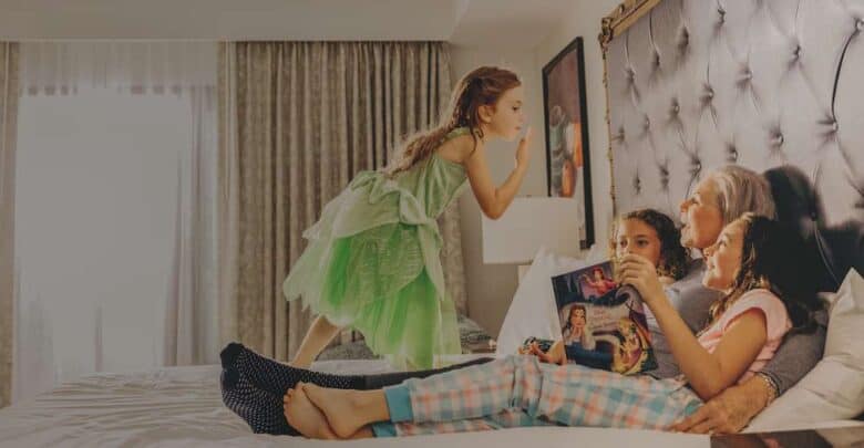 Three kids and their grandmother lounge on a bed, flipping through a Disney magazine in a sunlit, family-themed resort room.