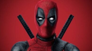 Deadpool, played by Ryan Reynolds, strikes a pose in his unmistakable red and black suit with twin swords strapped to his back, standing against a dramatic red backdrop. Marvel fans at the theme park are buzzing with excitement for his upcoming Avengers: Doomsday appearance.