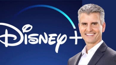 A sharply dressed Disney executive stands confidently before the glowing Disney+ logo on a vibrant blue backdrop, capturing the excitement and innovation fans associate with the new era of Disney entertainment and theme park experiences.